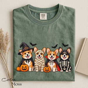 May include: A sage green t-shirt featuring a graphic of four Corgi dogs dressed in Halloween costumes: a witch with a pumpkin, a mummy, a vampire, and a skeleton. The shirt has a crew neckline and a Comfort Colors brand label at the inner neck.