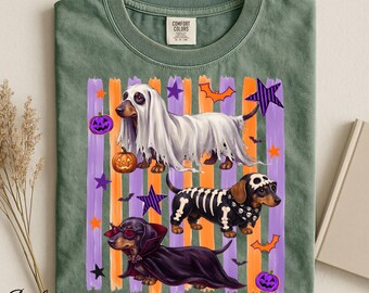 Op de afbeelding: Een saliegroen t-shirt met een Comfort Colors-label in de nek. De print toont drie teckels in Halloween-kostuums: een geest met een pompoen, een skelet en een vampier met een cape, tegen een achtergrond van oranje en paarse strepen.