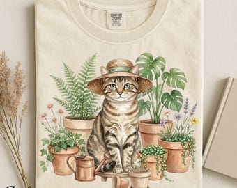 Op de afbeelding: Ivoor kleurig t-shirt met een aquarel illustratie van een tabby kat met een strohoed, omringd door potplanten en tuingereedschap. De tekst "Color Ivory" staat linksonder. Het shirt heeft een Comfort Colors label.