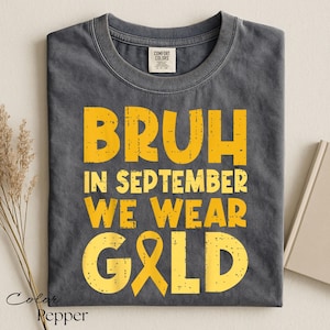 May include: A charcoal gray crew neck t-shirt with yellow distressed lettering that reads BRUH IN SEPTEMBER WE WEAR GOLD, featuring a gold awareness ribbon replacing the letter O in the word GOLD. The shirt has a Comfort Colors brand label.