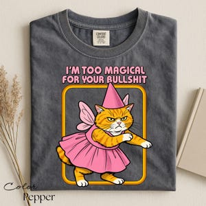 May include: A charcoal gray t-shirt featuring a graphic of an orange tabby cat wearing a pink fairy dress, wings, and a pointed hat. The text above the cat reads, I'M TOO MAGICAL FOR YOUR BULLSHIT, in pink letters inside a yellow-bordered frame.