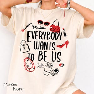 May include: Ivory-colored t-shirt with the phrase "EVERYBODY WANTS TO BE US" in black, along with illustrations of fashion accessories like sunglasses, a handbag, and high heels. The text "THE DEVIL WEARS PRADA" is also present.