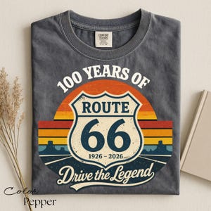May include: A dark grey t-shirt with a Route 66 graphic. The graphic features a shield with "Route 66" and "66" in the center, with the text "100 Years Of" above and "Drive the Legend" below. The shirt is labeled "Color Pepper".