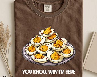 Op de afbeelding: Een bruin t-shirt met korte mouwen en een afbeelding van een bord met gevulde eieren met gele dooiers en bruine spikkels. Onder de illustratie staat de tekst "YOU KNOW WHY I'M HERE" in witte hoofdletters. Het shirt heeft een Comfort Colors label.