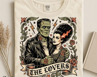 Op de afbeelding: Een ivoorkleurig t-shirt met een grafische illustratie van het monster van Frankenstein en zijn bruid. Het ontwerp bevat rode rozen, groene bladeren, bliksemschichten en een banner onderaan met de tekst THE LOVERS in zwarte letters.