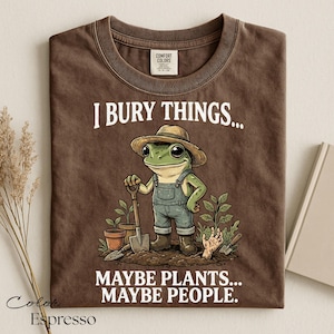 Może przedstawiać: Brązowy t-shirt z grafiką żaby w słomkowym kapeluszu i ogrodniczkach, trzymającej łopatę w ogrodzie. Napis głosi I BURY THINGS... MAYBE PLANTS... MAYBE PEOPLE. Koszulka posiada metkę Comfort Colors przy wewnętrznej stronie kołnierza.