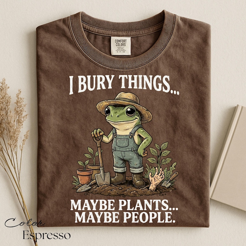 Może przedstawiać: Brązowy t-shirt z grafiką żaby w słomkowym kapeluszu i ogrodniczkach, trzymającej łopatę w ogrodzie. Napis głosi I BURY THINGS... MAYBE PLANTS... MAYBE PEOPLE. Koszulka posiada metkę Comfort Colors przy wewnętrznej stronie kołnierza.