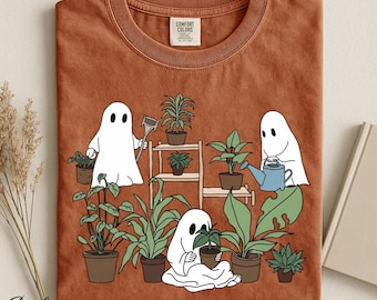 Op de afbeelding: Een roestkleurig t-shirt met een afbeelding van drie witte geesten die verschillende groene kamerplanten op een houten plank verzorgen. Eén geest houdt een schepje vast, een andere gebruikt een blauwe gieter en de derde houdt een plantje vast.