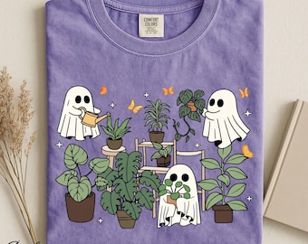 Op de afbeelding: Een violet t-shirt met een grafische print van drie witte geesten die verschillende groene potplanten op planken verzorgen. Het ontwerp bevat kleine oranje vlinders en witte steraccenten. Het shirt heeft een Comfort Colors label.