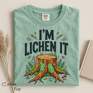 Puede incluir: Camiseta verde claro con el texto "I'M LICHEN IT" sobre una ilustraci&oacute;n de toc&oacute;n de &aacute;rbol. El dise&ntilde;o incluye hojas, setas y vegetaci&oacute;n. La camiseta est&aacute; doblada y tiene una etiqueta Comfort Colors.