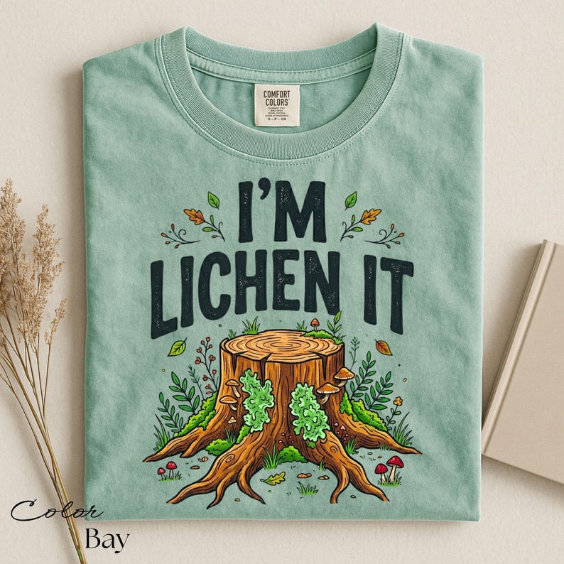 Puede incluir: Camiseta verde claro con el texto "I'M LICHEN IT" sobre una ilustraci&oacute;n de toc&oacute;n de &aacute;rbol. El dise&ntilde;o incluye hojas, setas y vegetaci&oacute;n. La camiseta est&aacute; doblada y tiene una etiqueta Comfort Colors.