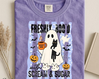 Op de afbeelding: Een violet t-shirt met een afbeelding van een geest die een zwarte kat en een oranje mok met de tekst SPOOKY BREW vasthoudt. Het ontwerp bevat pompoenen, vleermuizen, spinnenwebben en de tekst FRESHLY BOO'D with SCREAM & SUGAR in zwart.