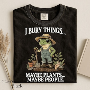 Może przedstawiać: Czarny t-shirt z grafiką żaby w słomkowym kapeluszu i dżinsowych ogrodniczkach, trzymającej łopatę w ogrodzie. Napis głosi I BURY THINGS... MAYBE PLANTS... MAYBE PEOPLE. Koszulka posiada okrągły dekolt oraz metkę marki Comfort Colors.
