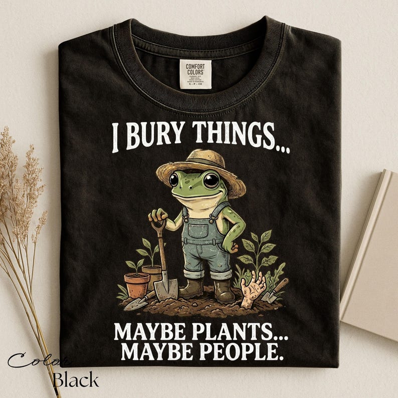 Może przedstawiać: Czarny t-shirt z grafiką żaby w słomkowym kapeluszu i dżinsowych ogrodniczkach, trzymającej łopatę w ogrodzie. Napis głosi I BURY THINGS... MAYBE PLANTS... MAYBE PEOPLE. Koszulka posiada okrągły dekolt oraz metkę marki Comfort Colors.
