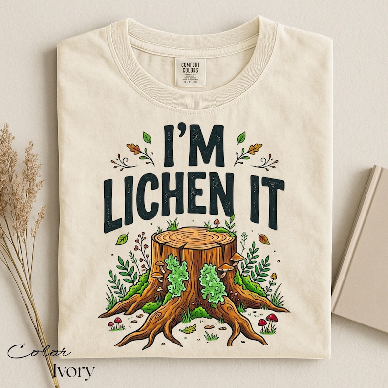 Puede incluir: Camiseta de color marfil con el texto "I'M LICHEN IT" sobre una ilustraci&oacute;n de un toc&oacute;n de &aacute;rbol. El toc&oacute;n presenta musgo verde, setas y follaje. La camiseta es de la marca Comfort Colors.