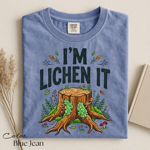 Puede incluir: Camiseta azul vaquero con el texto "I'M LICHEN IT" sobre una ilustraci&oacute;n detallada de un toc&oacute;n de &aacute;rbol con setas y follaje. La camiseta tiene cuello redondo y una etiqueta "Comfort Colors".