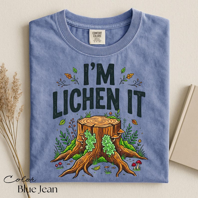 Puede incluir: Camiseta azul vaquero con el texto "I'M LICHEN IT" sobre una ilustraci&oacute;n detallada de un toc&oacute;n de &aacute;rbol con setas y follaje. La camiseta tiene cuello redondo y una etiqueta "Comfort Colors".