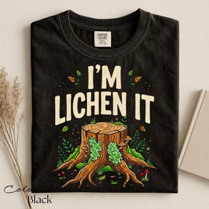 Puede incluir: Camiseta negra con el texto "I'M LICHEN IT" sobre un gr&aacute;fico de toc&oacute;n de &aacute;rbol con setas y vegetaci&oacute;n. La camiseta es de la marca Comfort Colors.