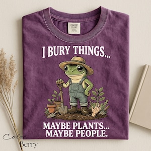 Może przedstawiać: Fioletowy t-shirt z grafiką żaby w słomkowym kapeluszu i ogrodniczkach, trzymającej łopatę w ogrodzie. Napis głosi I BURY THINGS... MAYBE PLANTS... MAYBE PEOPLE. Koszulka posiada metkę Comfort Colors przy wewnętrznej stronie kołnierza.