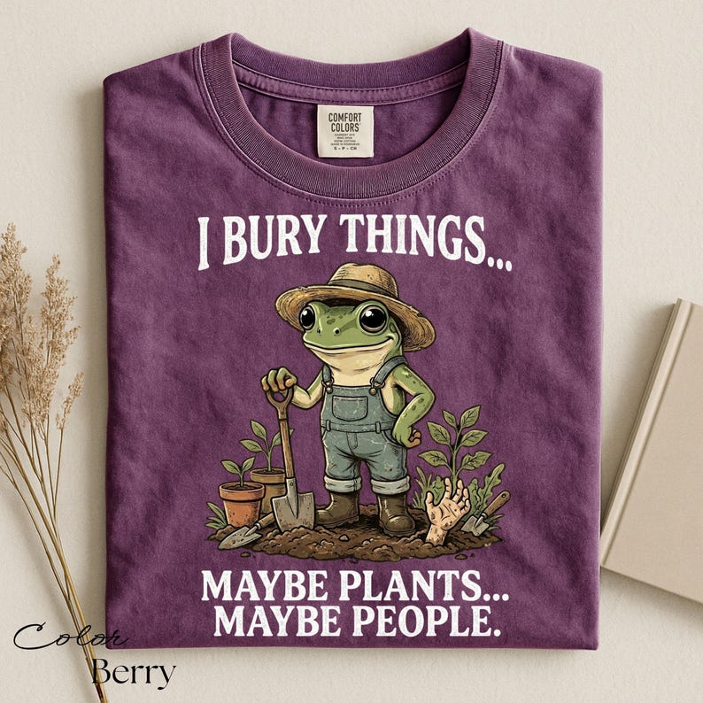 Może przedstawiać: Fioletowy t-shirt z grafiką żaby w słomkowym kapeluszu i ogrodniczkach, trzymającej łopatę w ogrodzie. Napis głosi I BURY THINGS... MAYBE PLANTS... MAYBE PEOPLE. Koszulka posiada metkę Comfort Colors przy wewnętrznej stronie kołnierza.