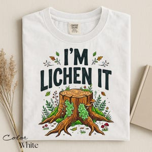 Puede incluir: Camiseta blanca con el texto "I'M LICHEN IT" sobre una ilustraci&oacute;n de un toc&oacute;n de &aacute;rbol. El gr&aacute;fico presenta un toc&oacute;n de &aacute;rbol con musgo verde, setas y follaje. La camiseta es de la marca Comfort Colors.