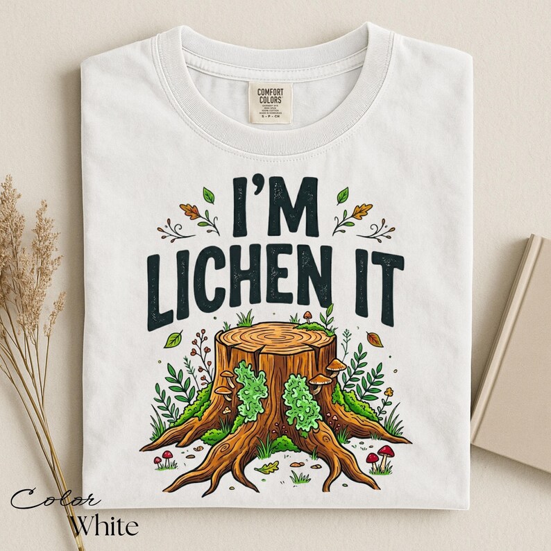 Puede incluir: Camiseta blanca con el texto "I'M LICHEN IT" sobre una ilustraci&oacute;n de un toc&oacute;n de &aacute;rbol. El gr&aacute;fico presenta un toc&oacute;n de &aacute;rbol con musgo verde, setas y follaje. La camiseta es de la marca Comfort Colors.