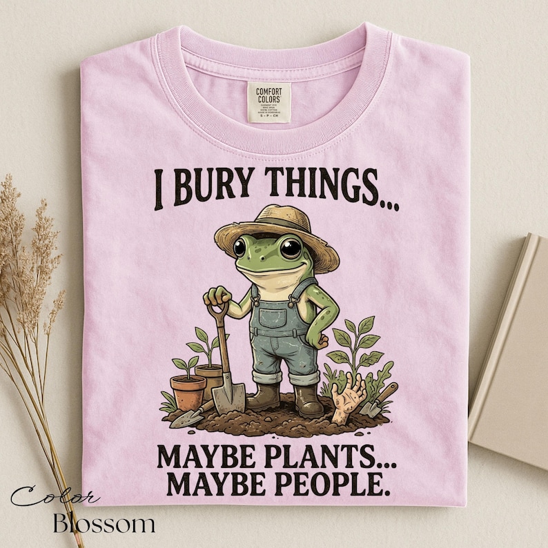 Może przedstawiać: Jasnor&oacute;żowy t-shirt z grafiką żaby w słomkowym kapeluszu i ogrodniczkach, trzymającej łopatę w ziemi. Napis głosi I BURY THINGS... MAYBE PLANTS... MAYBE PEOPLE. Koszulka posiada metkę marki Comfort Colors przy kołnierzyku.