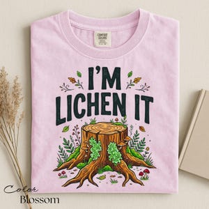 Puede incluir: Camiseta rosa claro con la frase "I'M LICHEN IT" en negro. El dise&ntilde;o presenta un toc&oacute;n de &aacute;rbol con setas, musgo y follaje. Marca Comfort Colors.