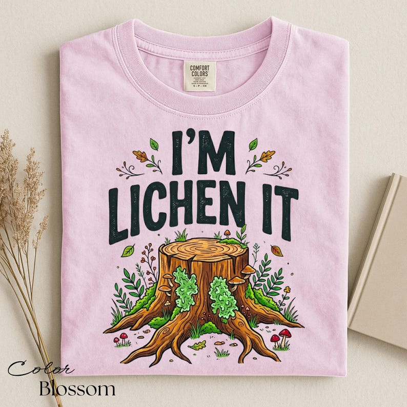 Puede incluir: Camiseta rosa claro con la frase "I'M LICHEN IT" en negro. El dise&ntilde;o presenta un toc&oacute;n de &aacute;rbol con setas, musgo y follaje. Marca Comfort Colors.