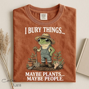 Może przedstawiać: T-shirt w kolorze rdzawym z grafiką żaby w słomkowym kapeluszu i ogrodniczkach, trzymającej łopatę. Tekst głosi: I BURY THINGS... MAYBE PLANTS... MAYBE PEOPLE. Wz&oacute;r zawiera rośliny doniczkowe oraz dłoń wyłaniającą się z ziemi.