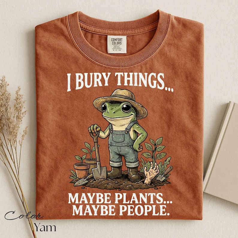 Może przedstawiać: T-shirt w kolorze rdzawym z grafiką żaby w słomkowym kapeluszu i ogrodniczkach, trzymającej łopatę. Tekst głosi: I BURY THINGS... MAYBE PLANTS... MAYBE PEOPLE. Wz&oacute;r zawiera rośliny doniczkowe oraz dłoń wyłaniającą się z ziemi.