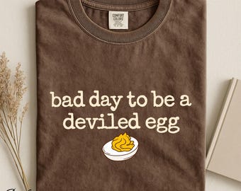 Op de afbeelding: Een bruin t-shirt met korte mouwen en een ronde hals. Het shirt heeft de witte tekst "bad day to be a deviled egg" gedrukt boven een afbeelding van een gehalveerd gevuld ei met een gele dooier en wit eiwit. Het merklabel is Comfort Colors.