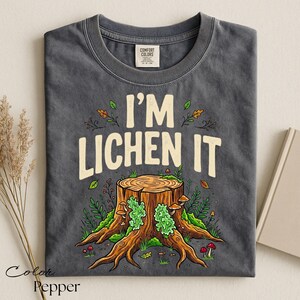 Puede incluir: Camiseta gris oscuro con el texto "I'M LICHEN IT" sobre una ilustraci&oacute;n de toc&oacute;n de &aacute;rbol. El toc&oacute;n es marr&oacute;n con musgo verde y setas. La camiseta es de material Comfort Colors.