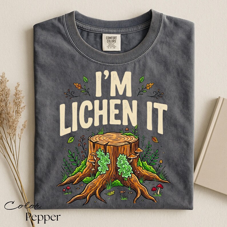Puede incluir: Camiseta gris oscuro con el texto "I'M LICHEN IT" sobre una ilustraci&oacute;n de toc&oacute;n de &aacute;rbol. El toc&oacute;n es marr&oacute;n con musgo verde y setas. La camiseta es de material Comfort Colors.