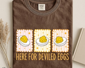 Op de afbeelding: Een bruin t-shirt met korte mouwen voorzien van drie rechthoekige kaders met roze en gele verticale strepen. Elk kader bevat een afbeelding van een gevuld ei. De tekst "HERE FOR DEVILED EGGS" staat in het geel onder de afbeeldingen.