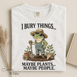 Może przedstawiać: Biały t-shirt z grafiką zielonej żaby w słomkowym kapeluszu i dżinsowych ogrodniczkach, trzymającej łopatę w ogrodzie. Tekst brzmi I BURY THINGS... MAYBE PLANTS... MAYBE PEOPLE. Wz&oacute;r zawiera rośliny doniczkowe oraz wystającą z ziemi dłoń.