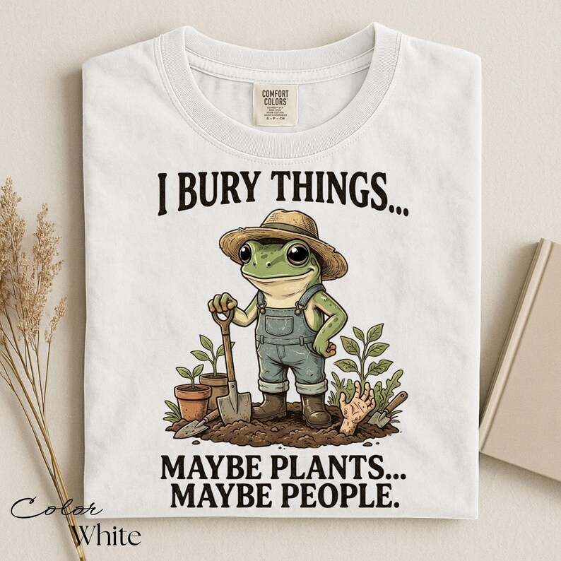 Może przedstawiać: Biały t-shirt z grafiką zielonej żaby w słomkowym kapeluszu i dżinsowych ogrodniczkach, trzymającej łopatę w ogrodzie. Tekst brzmi I BURY THINGS... MAYBE PLANTS... MAYBE PEOPLE. Wz&oacute;r zawiera rośliny doniczkowe oraz wystającą z ziemi dłoń.