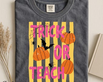 Op de afbeelding: Een antracietgrijs t-shirt met een afbeelding van verticale gele strepen, drie oranje pompoenen, een zwarte vleermuis en spinnenwebben. De tekst TRICK OR TEACH staat in dikke roze letters. Het shirt heeft een Comfort Colors label in de nek.