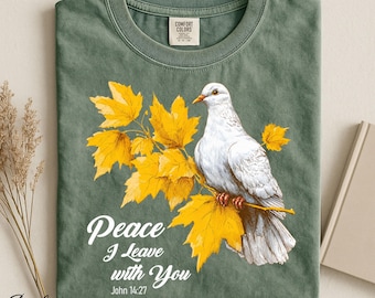 Op de afbeelding: Een saliegroen t-shirt met een witte duif zittend op een tak met felgele esdoornbladeren. De tekst "Peace I Leave with You" en "John 14:27" is in het wit onder de illustratie gedrukt. Het t-shirt heeft een ronde hals en een label.