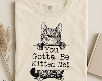 Op de afbeelding: Een ivoorkleurig t-shirt met een zwarte grafische illustratie van een kat die over een bordje kijkt. De tekst op het bordje luidt "You Gotta Be Kitten Me!". Het t-shirt heeft een ronde hals en een Comfort Colors label in de binnenkant van de kraag.