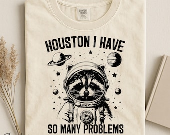 Op de afbeelding: Een ivoorkleurig t-shirt met een zwarte afbeelding van een wasbeer in een ruimtehelm. De tekst luidt HOUSTON I HAVE SO MANY PROBLEMS. Het ontwerp bevat illustraties van planeten en sterren rondom de wasbeer. Het shirt heeft een Comfort Colors label.