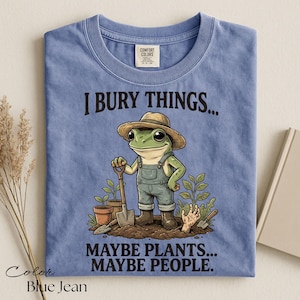 Może przedstawiać: Dżinsowoniebieski t-shirt z grafiką żaby w słomkowym kapeluszu i ogrodniczkach, trzymającej łopatę. Napis głosi I BURY THINGS... MAYBE PLANTS... MAYBE PEOPLE. Wz&oacute;r zawiera rośliny doniczkowe oraz dłoń wyłaniającą się z ziemi.