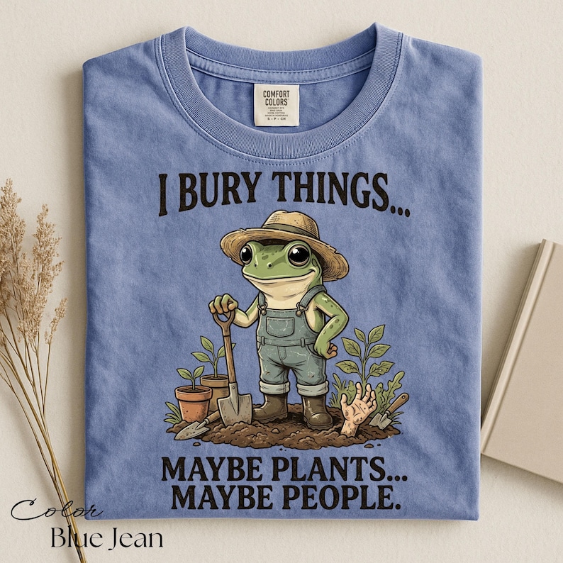 Może przedstawiać: Dżinsowoniebieski t-shirt z grafiką żaby w słomkowym kapeluszu i ogrodniczkach, trzymającej łopatę. Napis głosi I BURY THINGS... MAYBE PLANTS... MAYBE PEOPLE. Wz&oacute;r zawiera rośliny doniczkowe oraz dłoń wyłaniającą się z ziemi.