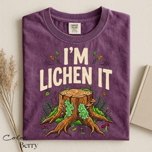 Puede incluir: Camiseta de color bayas con el texto "I'M LICHEN IT" sobre una ilustraci&oacute;n de toc&oacute;n de &aacute;rbol. El gr&aacute;fico presenta un toc&oacute;n de &aacute;rbol detallado con musgo verde, setas y follaje. La camiseta es de material Comfort Colors.