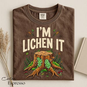 Puede incluir: Camiseta color espresso con el texto "I'M LICHEN IT" sobre una ilustraci&oacute;n detallada de un toc&oacute;n de &aacute;rbol cubierto de l&iacute;quenes verdes, setas y follaje. La camiseta est&aacute; hecha de material Comfort Colors.