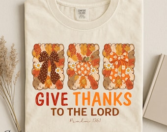 Op de afbeelding: Een ivoorkleurig t-shirt met drie rechthoekige panelen met herfstbladeren, madeliefjes, een strik met stippen, een pompoen en een bloemrijke strik. Onder de afbeeldingen staat "GIVE THANKS TO THE LORD" in oranje en bruin, gevolgd door "Psalm 136:1".