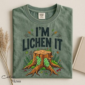 Puede incluir: Camiseta verde musgo con el texto "I'M LICHEN IT" sobre una ilustraci&oacute;n de un toc&oacute;n de &aacute;rbol. La ilustraci&oacute;n presenta un toc&oacute;n de &aacute;rbol con musgo verde, setas y follaje. La camiseta es de la marca Comfort Colors.