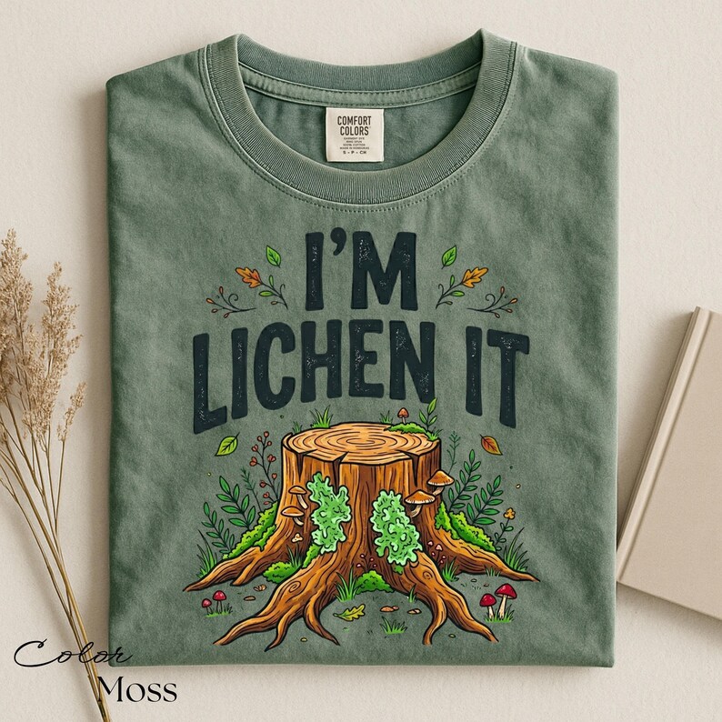 Puede incluir: Camiseta verde musgo con el texto "I'M LICHEN IT" sobre una ilustraci&oacute;n de un toc&oacute;n de &aacute;rbol. La ilustraci&oacute;n presenta un toc&oacute;n de &aacute;rbol con musgo verde, setas y follaje. La camiseta es de la marca Comfort Colors.