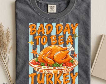 Op de afbeelding: Een antracietgrijs t-shirt met ronde hals en een grafische print van een gebraden kalkoen op een schaal, omringd door herfstbladeren en bessen. De tekst BAD DAY TO BE A TURKEY staat in oranje letters, geaccentueerd door sterren en lichtblauwe strepen.
