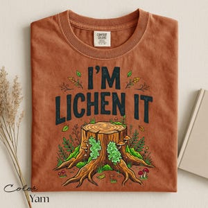Puede incluir: Una camiseta de color &oacute;xido con la frase "I'M LICHEN IT" en letras negras. El gr&aacute;fico presenta un toc&oacute;n de &aacute;rbol con l&iacute;quenes verdes, setas y follaje. La camiseta est&aacute; doblada y tiene una etiqueta en el centro superior.